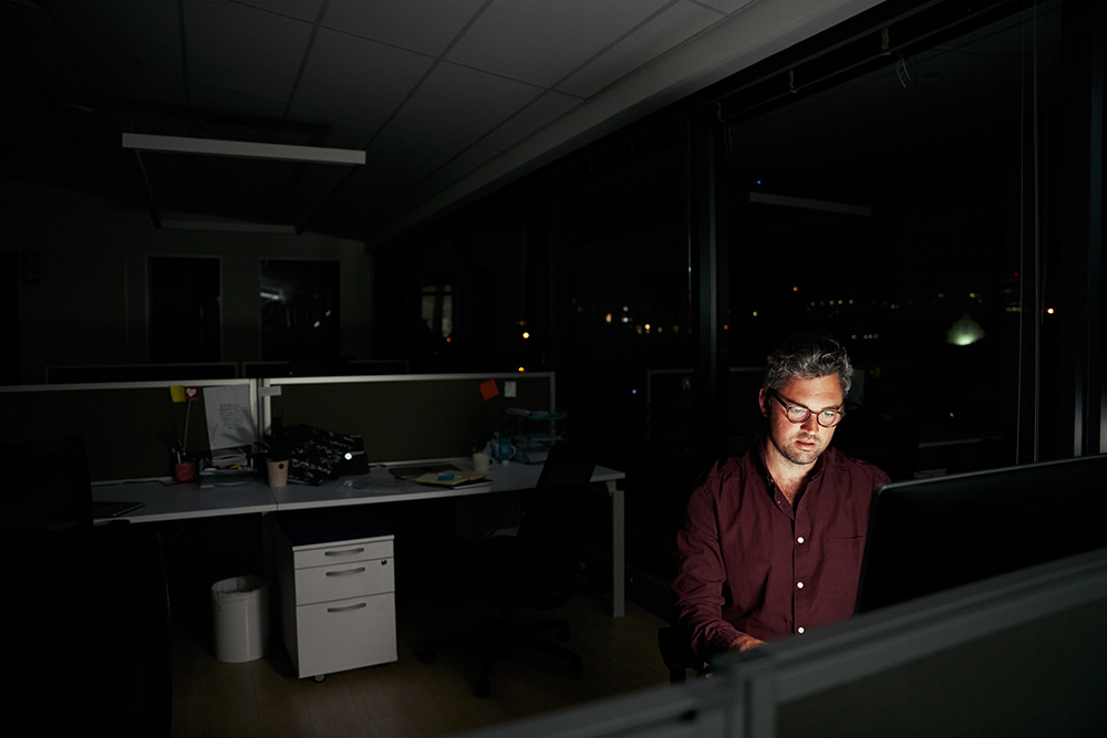 Middle aged businessman is sitting at a laptop in a dark office working on new deadline project – young male working late at night in the office